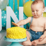 1 year old boy celebrating his first birthday with a yellow birthday cake and blue balloons in the background