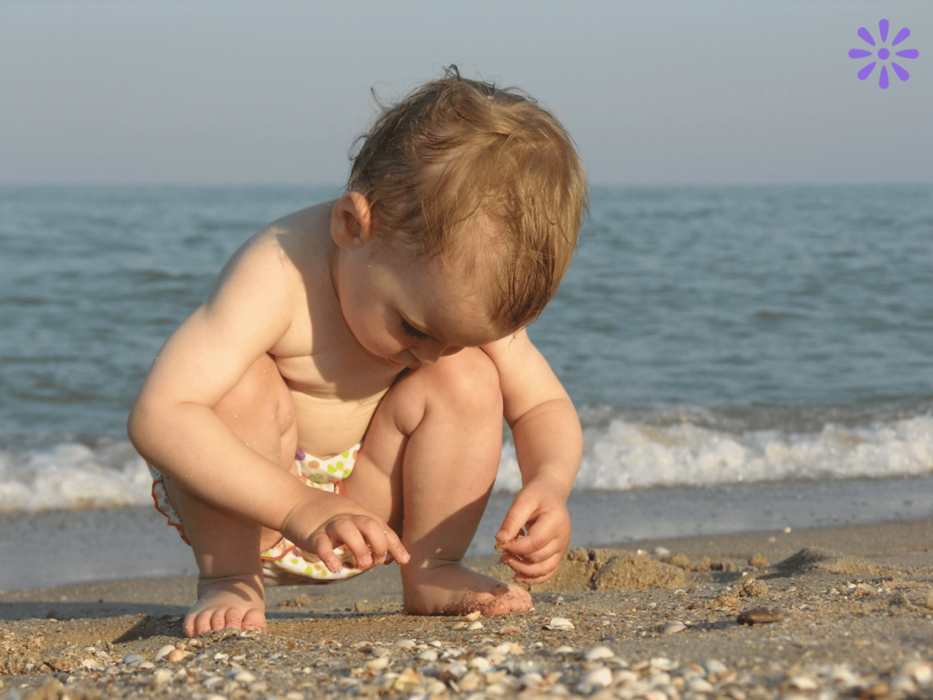 baby in a swim diaper looking at shells on the beach with the ocean in the background - beach hack for babies - learning about shells and sea life