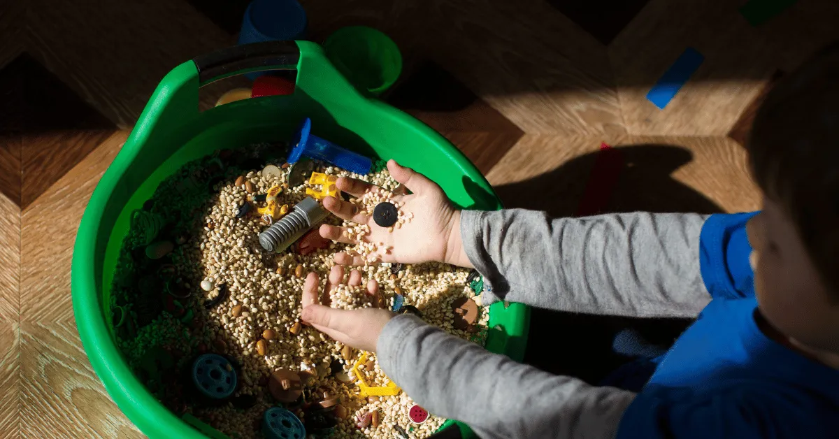 Child playing in a basket of grains with plastic shapes put together into a sensory basket